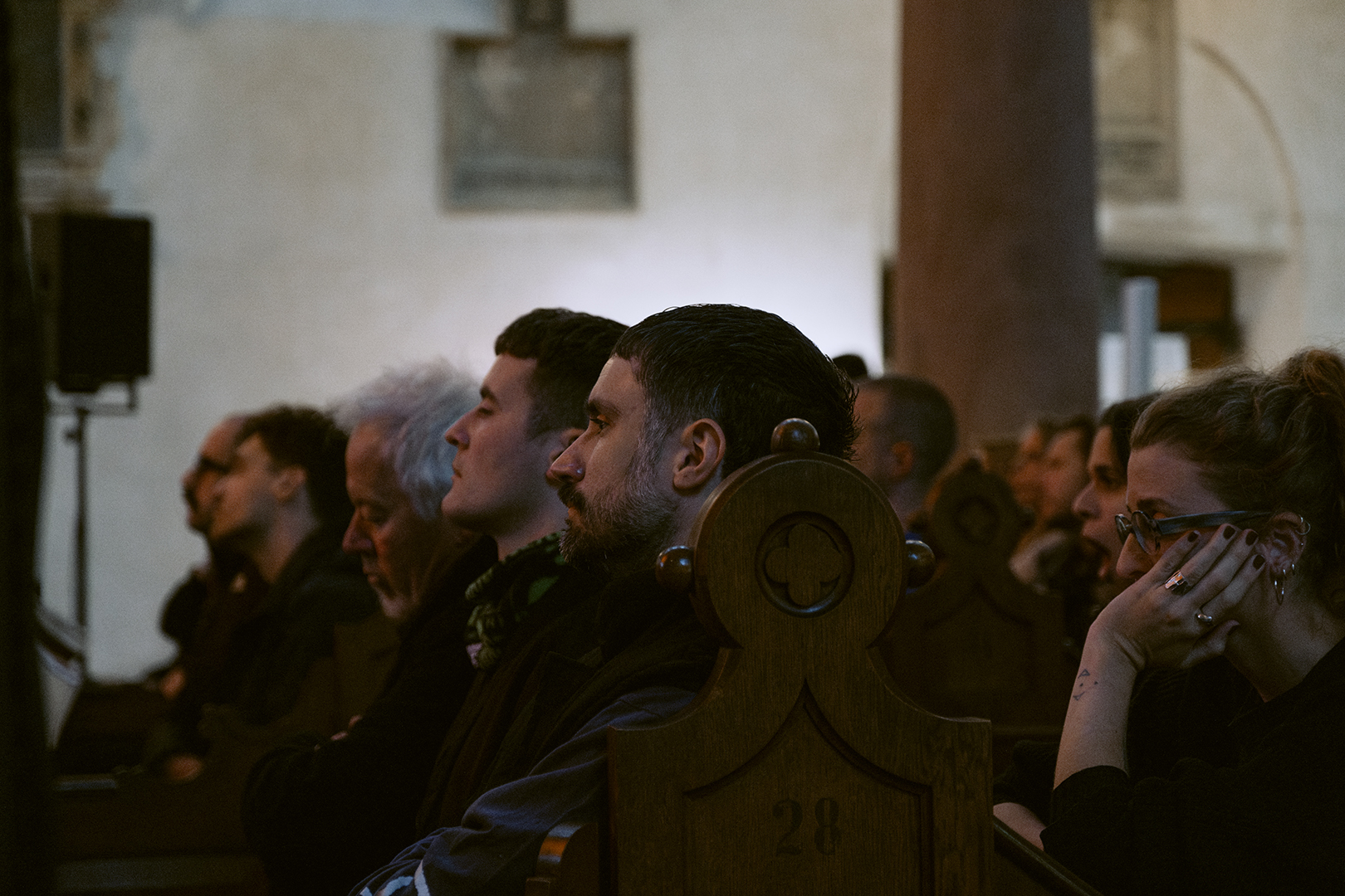 A photo of people sitting on church benches. They are all looking into the same direction, listening to the performance on stage.